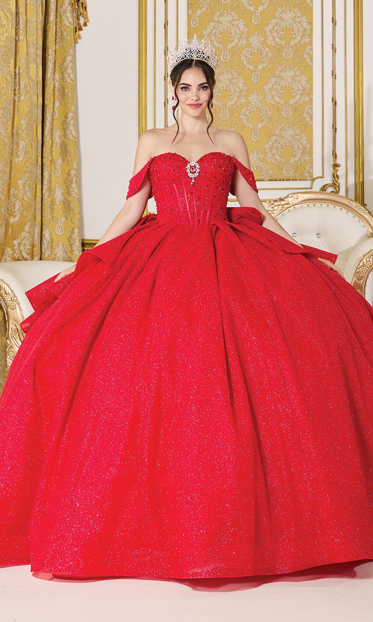 A young woman wears the Quinceanera Dress 1937 by Dancing Queen, featuring off-the-shoulder straps and sparkling red fabric, with a tiara and necklace, standing in an ornate gold and white room.